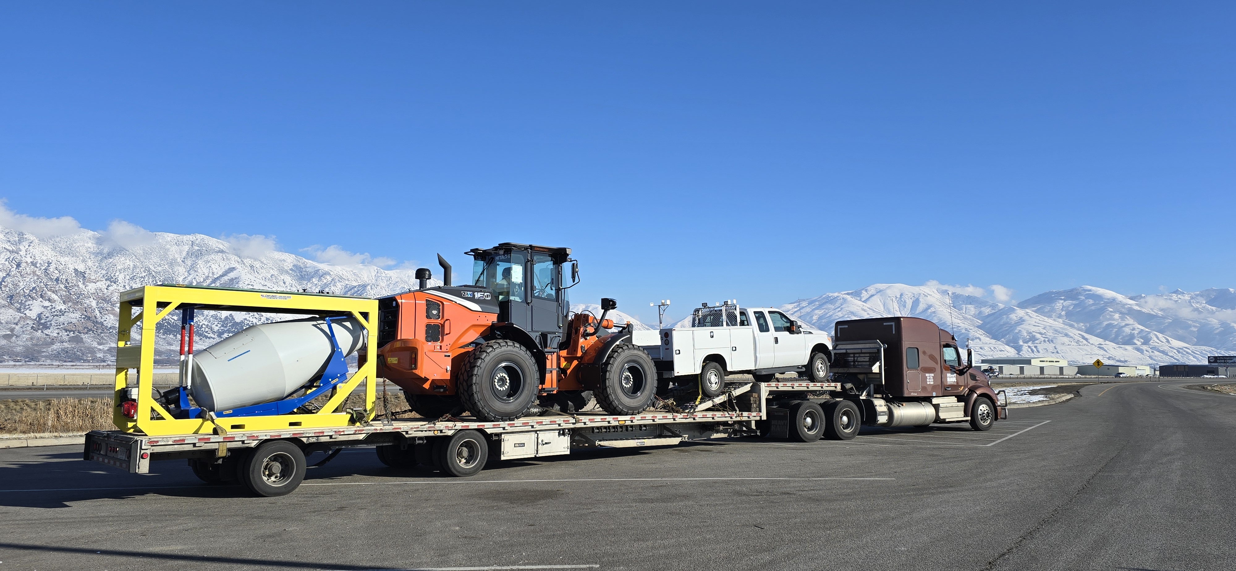 An HHS step-deck load chained down for transport: a wheel loader, a service pickup and a concrete mixer, snow-capped mountains behind