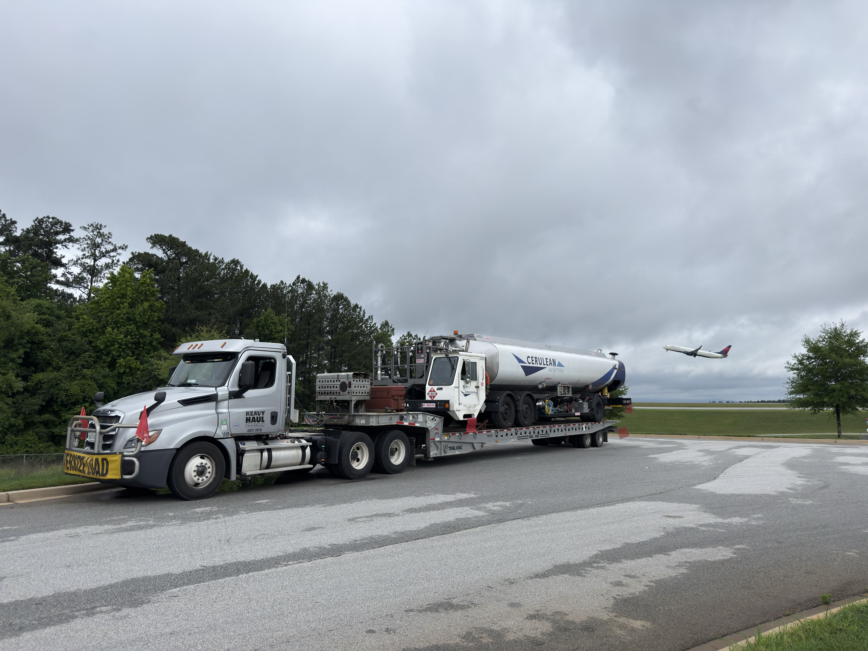 A white HHS day cab with an oversize load banner hauling an aviation refueling truck on a step-deck trailer, an airliner taking off behind