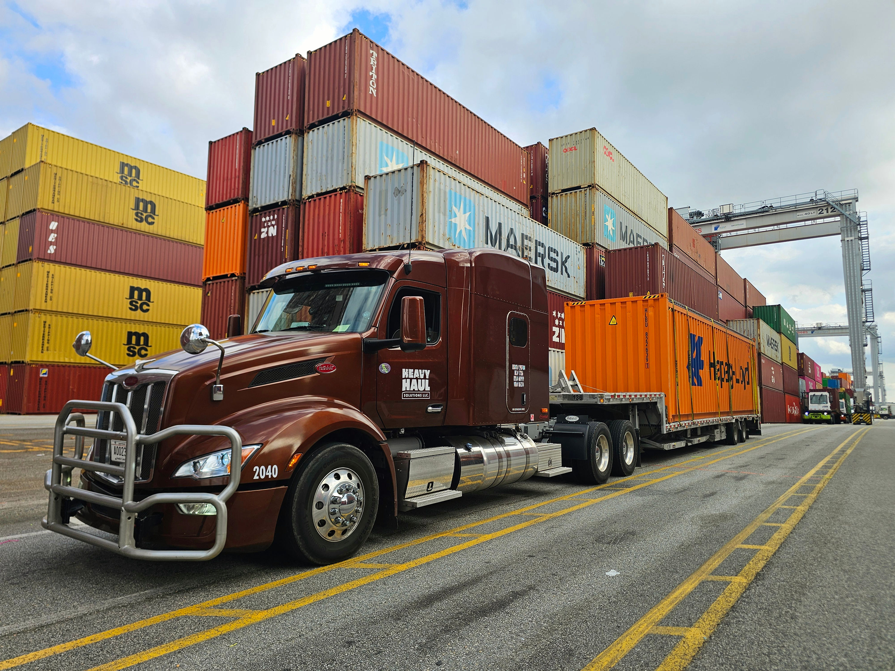 Maroon HHS Peterbilt hauling an orange container on a stepdeck through a container terminal
