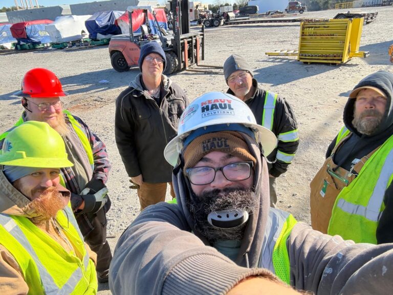 Group of Heavy Haul Solutions workers in safety vests and hard hats gathered at a jobsite with equipment and trailers in the background.