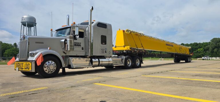 Semi tractor with yellow step deck trailer parked in an industrial yard, with water tower visible in background.