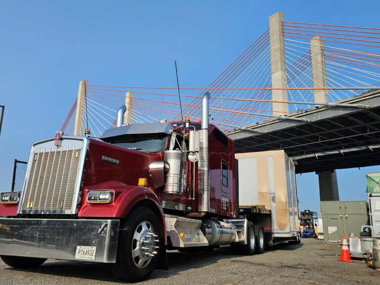 Semi tractor with enclosed trailer parked at industrial yard with cable-stayed bridge in background.