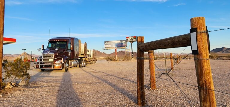 Semi tractor with stepdeck trailer parked at a rural yard with wooden fence gate and desert landscape in background.