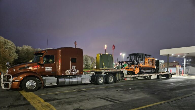 Heavy Haul Solutions semi truck with stepdeck trailer carrying an excavator and compactor, parked at night near a fuel station.