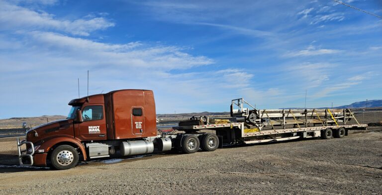 Semi tractor with stepdeck trailer carrying large industrial equipment and machinery, parked in an open yard under blue sky.