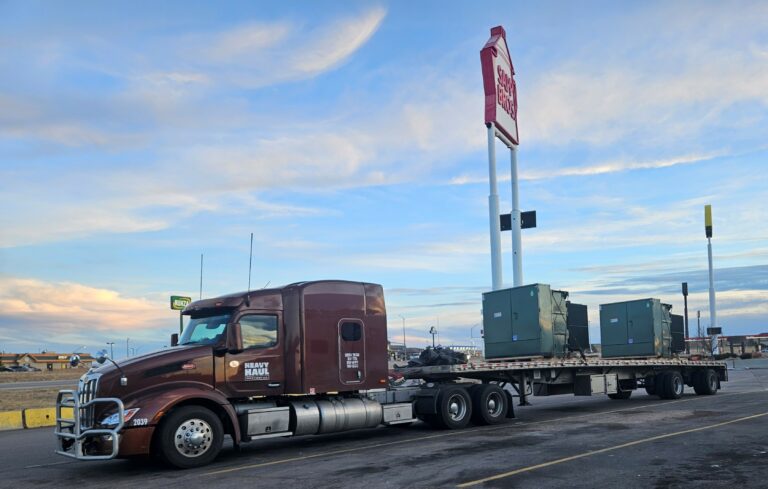 Heavy Haul Solutions semi tractor with sleeper cab and flatbed trailer loaded with large green industrial transformers parked at a truck stop.
