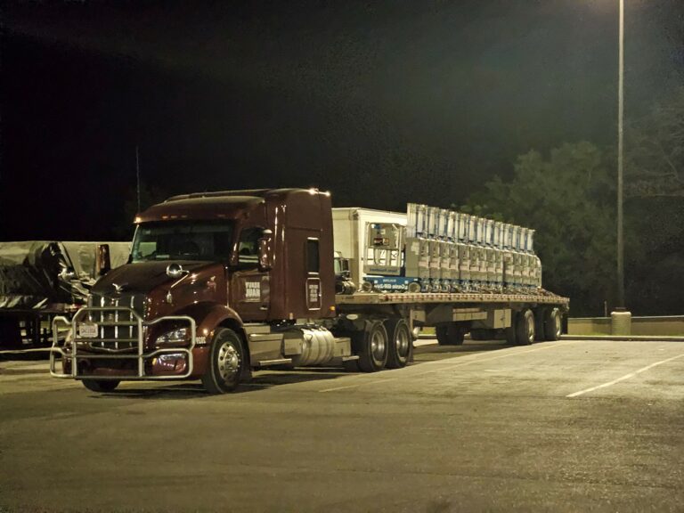 Semi tractor with flatbed trailer carrying stacked industrial units parked at night in a yard under lights.