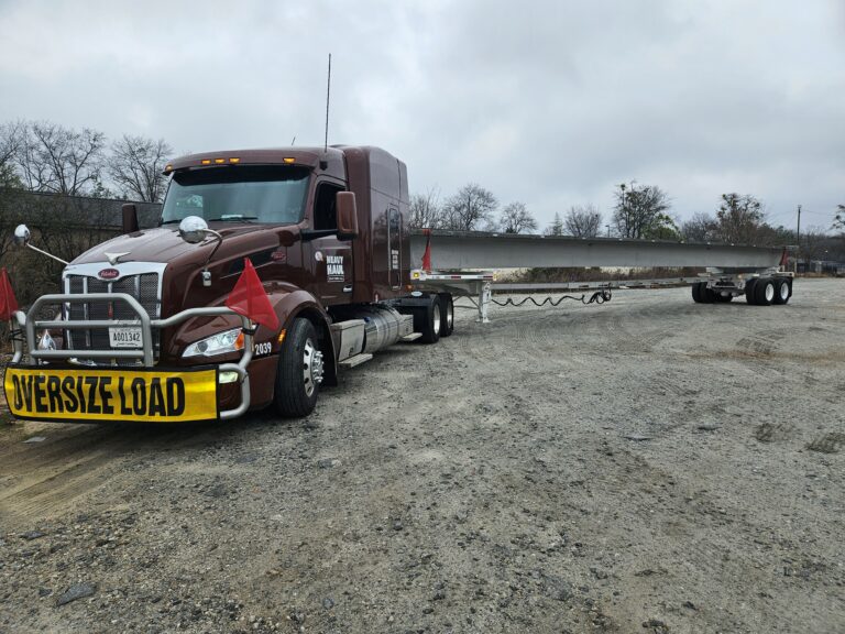 Semi tractor with oversize load sign hauling long industrial cargo on gravel yard.
