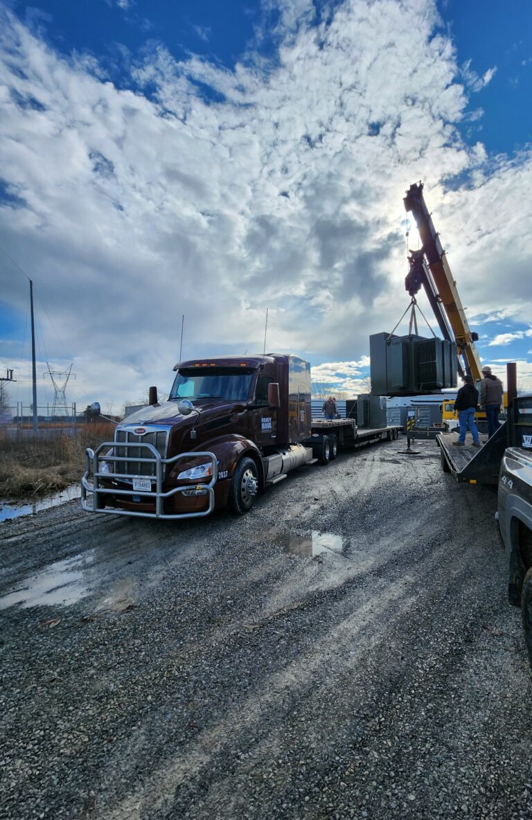 Semi tractor and stepdeck trailer parked at an industrial yard with crane and workers in the background under cloudy sky.