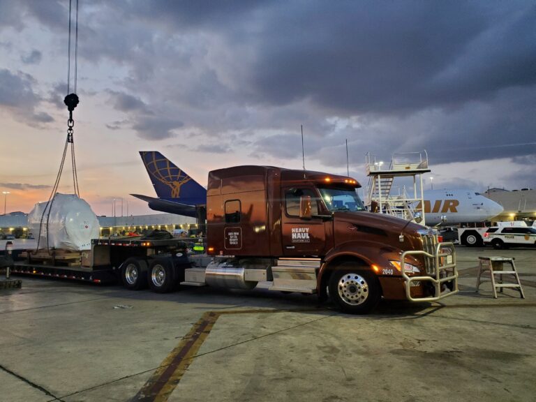 Heavy Haul Solutions semi truck with lowboy trailer parked at industrial facility, with aircraft and cargo equipment visible in background at dusk.