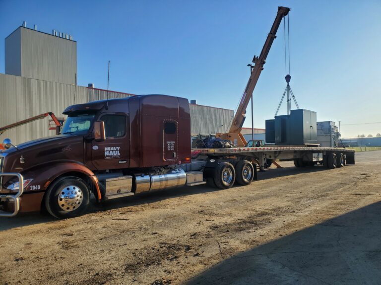 Heavy Haul Solutions semi truck with flatbed trailer parked at industrial yard, crane lifting large industrial unit onto trailer.