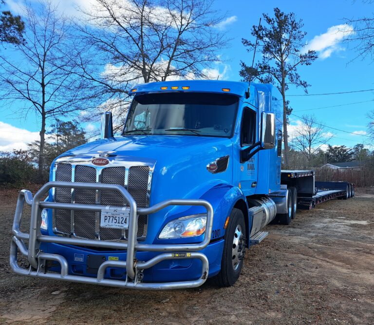 Blue semi tractor with brush guard parked at a dirt lot, pulling a dark RGN trailer with a smaller unit visible behind it.