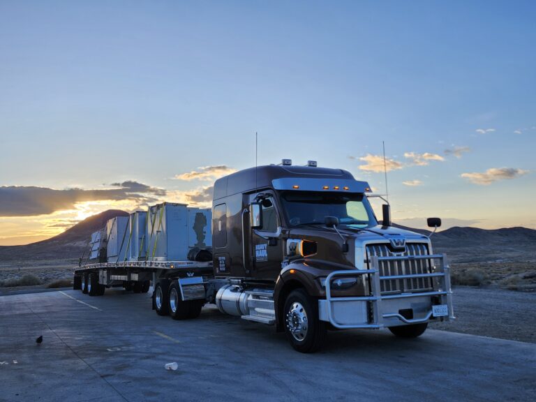 Semi tractor with flatbed trailer parked in desert landscape at sunset with mountains in background.