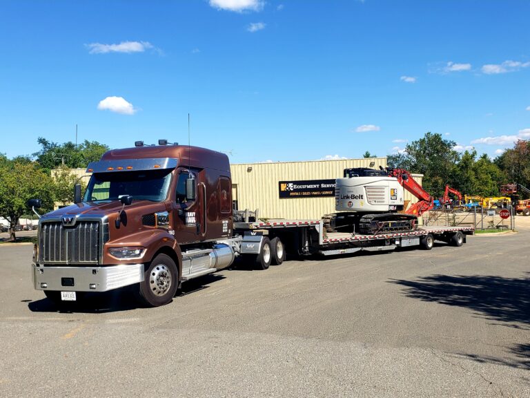 Semi tractor with stepdeck  trailer carrying construction equipment parked at a company yard with industrial buildings in background