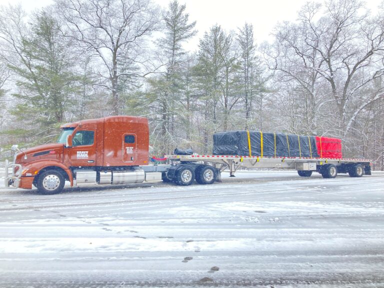 Semi tractor with flatbed trailer carrying wrapped industrial cargo parked in a snow lot with bare trees in background