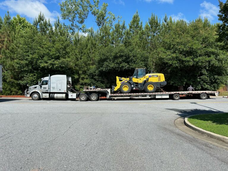 Semi truck with stepdeck trailer carrying yellow wheel loader, parked in lot with trees in background.