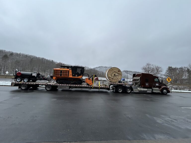 stepdeck trailer carrying orange construction equipment and a large wooden spool, parked at a rest area with bare trees and overcast sky in background.