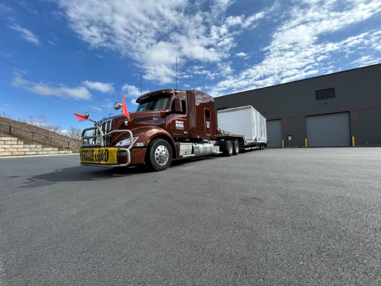 Semi tractor with stepdeck trailer parked in industrial yard with warehouse building and blue sky.