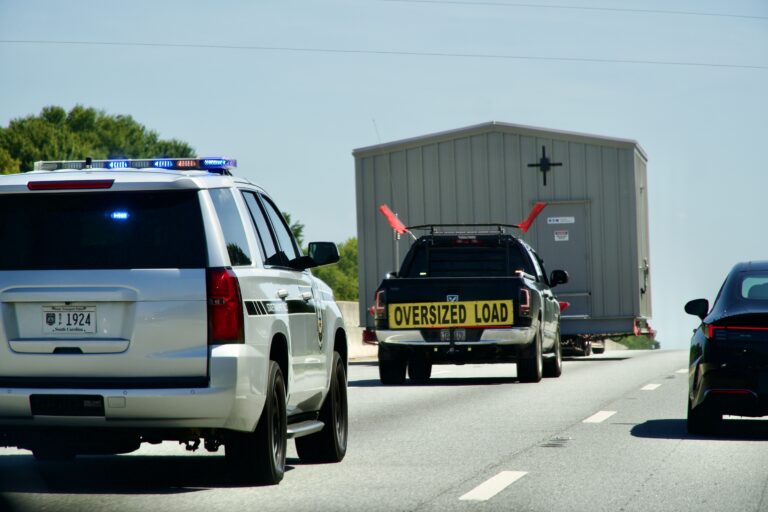 Police escort vehicle accompanying a truck marked 'OVERSIZED LOAD' on a highway with a large structure on an oversized trailer visible.