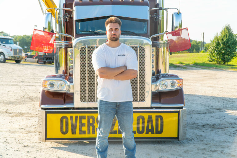 Truck driver in white t-shirt stands with arms crossed in front of a brown and tan semi tractor with yellow oversize load sign.
