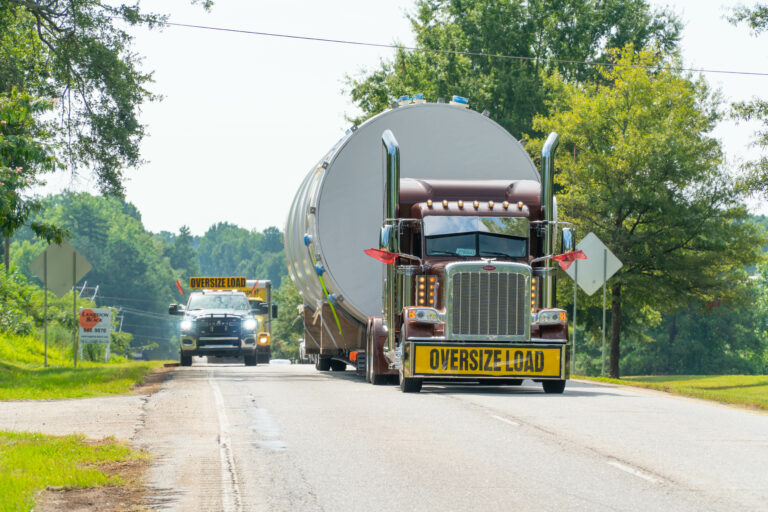 Semi truck with oversized tanker trailer marked with oversize load signs travels on rural highway with escort vehicle ahead.