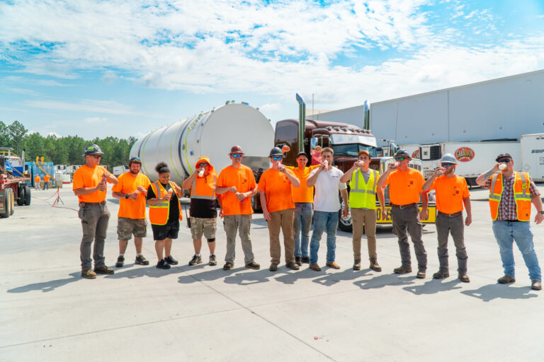 Team members in orange shirts stand in a line at a terminal yard in front of a tanker truck and large warehouse building.