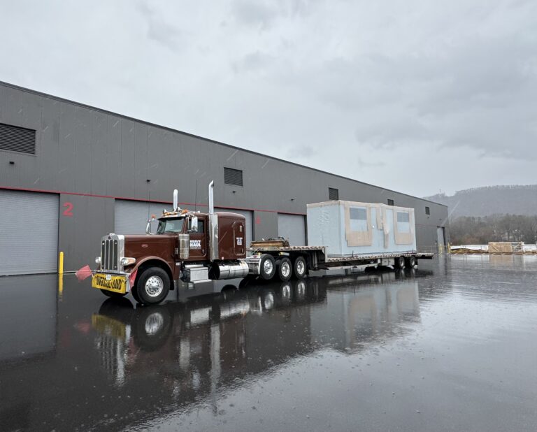 Semi tractor with stepdeck trailer carrying a white modular building unit parked outside an industrial facility in wet conditions.