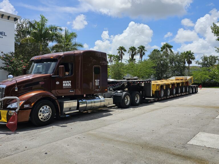 Semi tractor with Heavy Haul Solutions branding parked beside yellow step deck trailer in yard with palm trees under blue sky