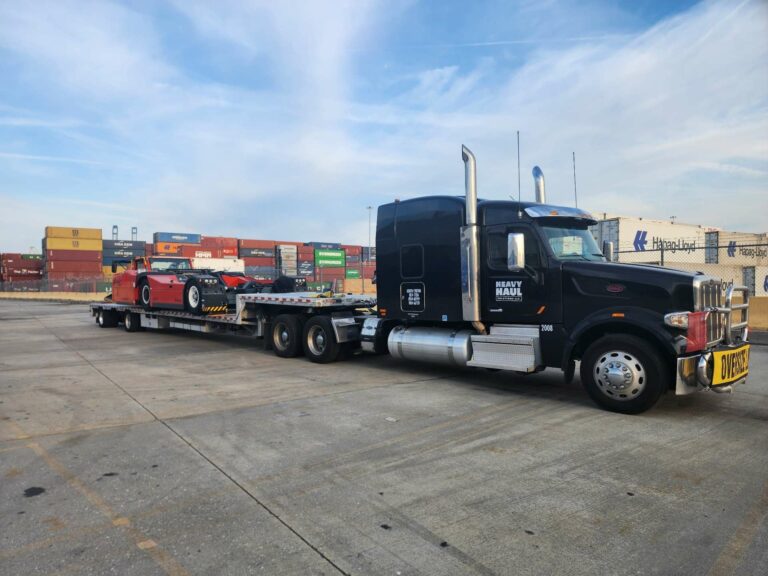 Heavy Haul Solutions semi tractor with chrome exhaust stacks parked at a terminal, towing a stepdeck trailer loaded with red machinery, shipping containers visible in background.