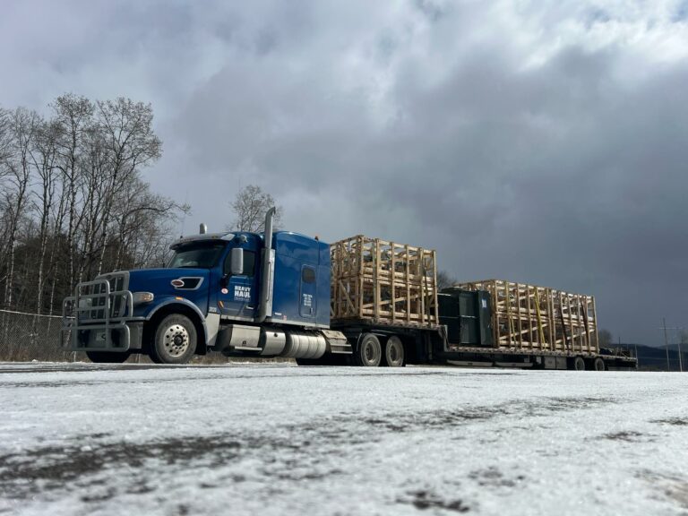 Semi tractor with stepdeck trailers loaded with wooden crates parked in an industrial lot under overcast sky.