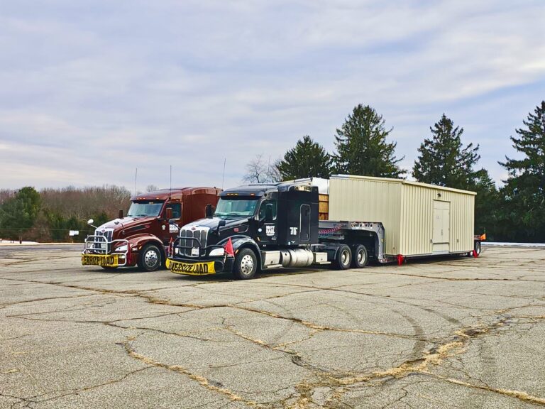 Two semi tractors with a large white enclosed cargo trailer parked in a paved yard with evergreen trees in the background.