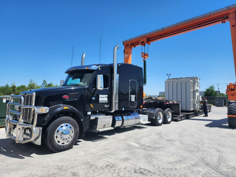 Semi tractor with dual sleeper cab parked at a terminal, carrying a large industrial module on a RGN trailer. Overhead gantry crane visible nearby.
