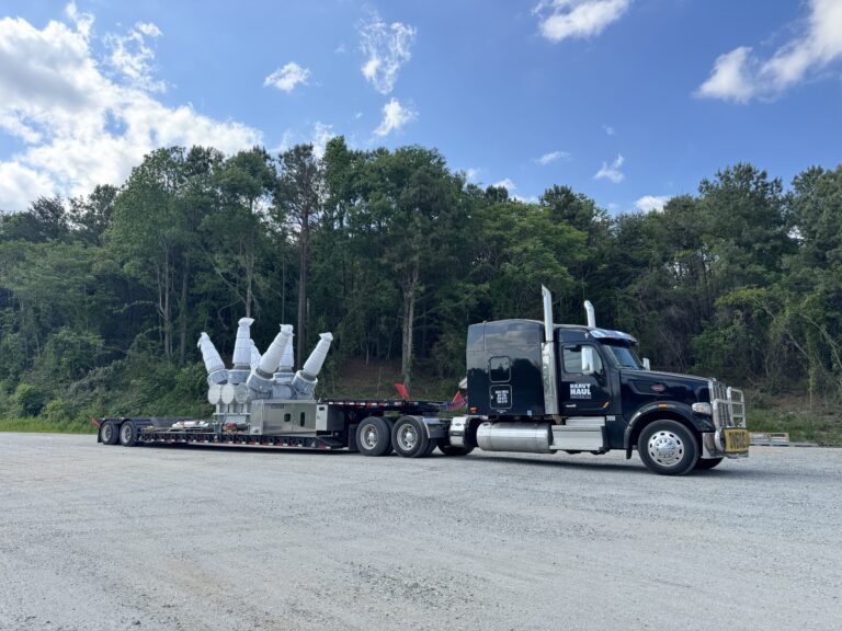 Semi truck with Heavy Haul Solutions branding parked alongside a RGN trailer carrying large white industrial transformer units, with forest in background.