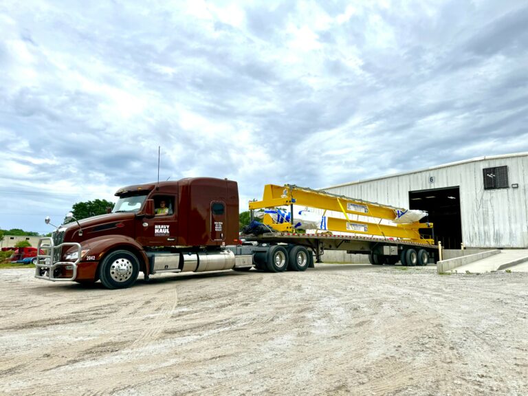 Semi tractor with Heavy Haul Solutions branding parked next to a yellow lowboy trailer carrying industrial machinery, with a metal warehouse building in background.