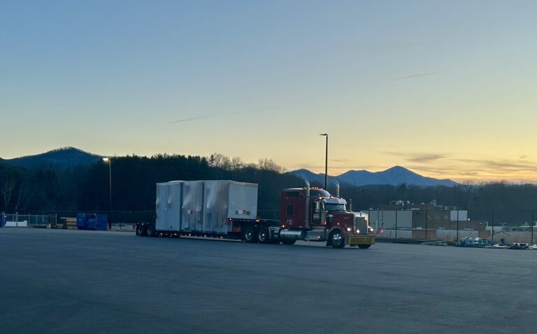 Semi tractor with enclosed white trailer parked in industrial yard at dusk, mountains visible in background.