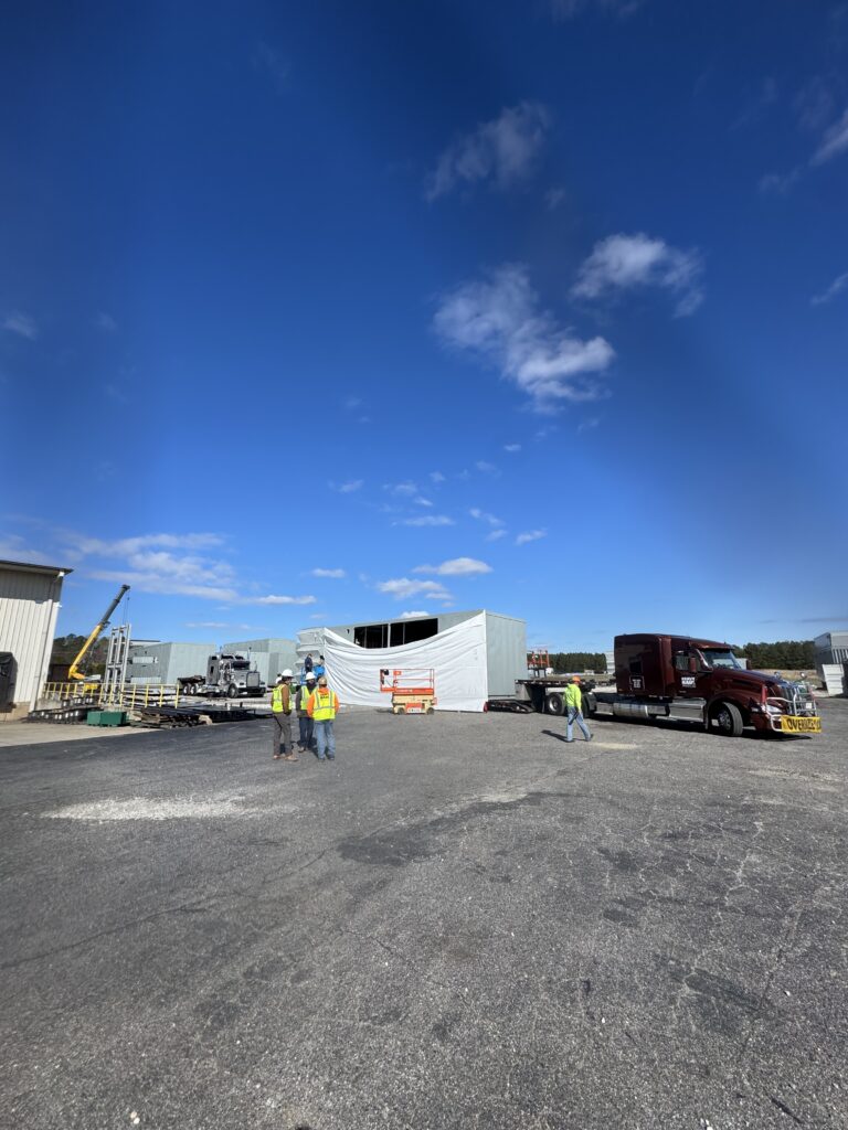 Trucking yard with RGN trailer covered in white tarp, semi truck, workers in safety vests, and industrial equipment under clear blue sky.