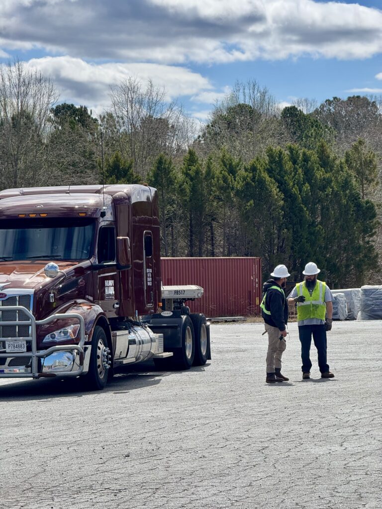 Tractor-trailer parked at yard with two workers in hard hats and safety vests standing nearby. Shipping container and forest visible in background.