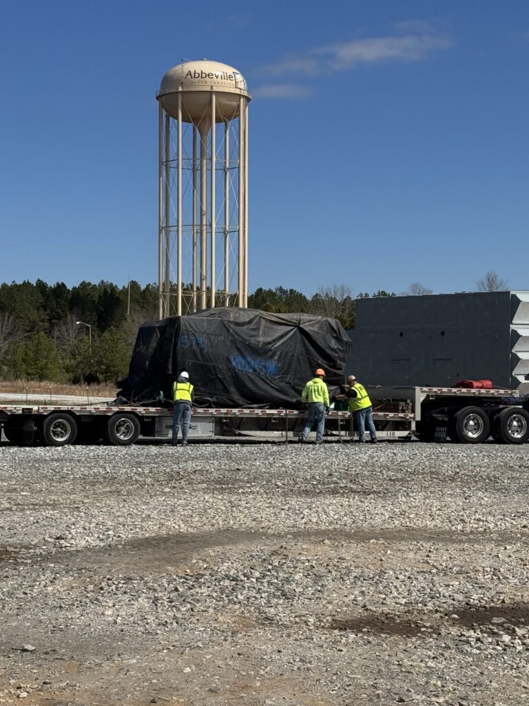 Stepdeck trailer parked at a gravel yard with a large covered industrial unit, water tower visible in background, three workers in safety vests nearby.