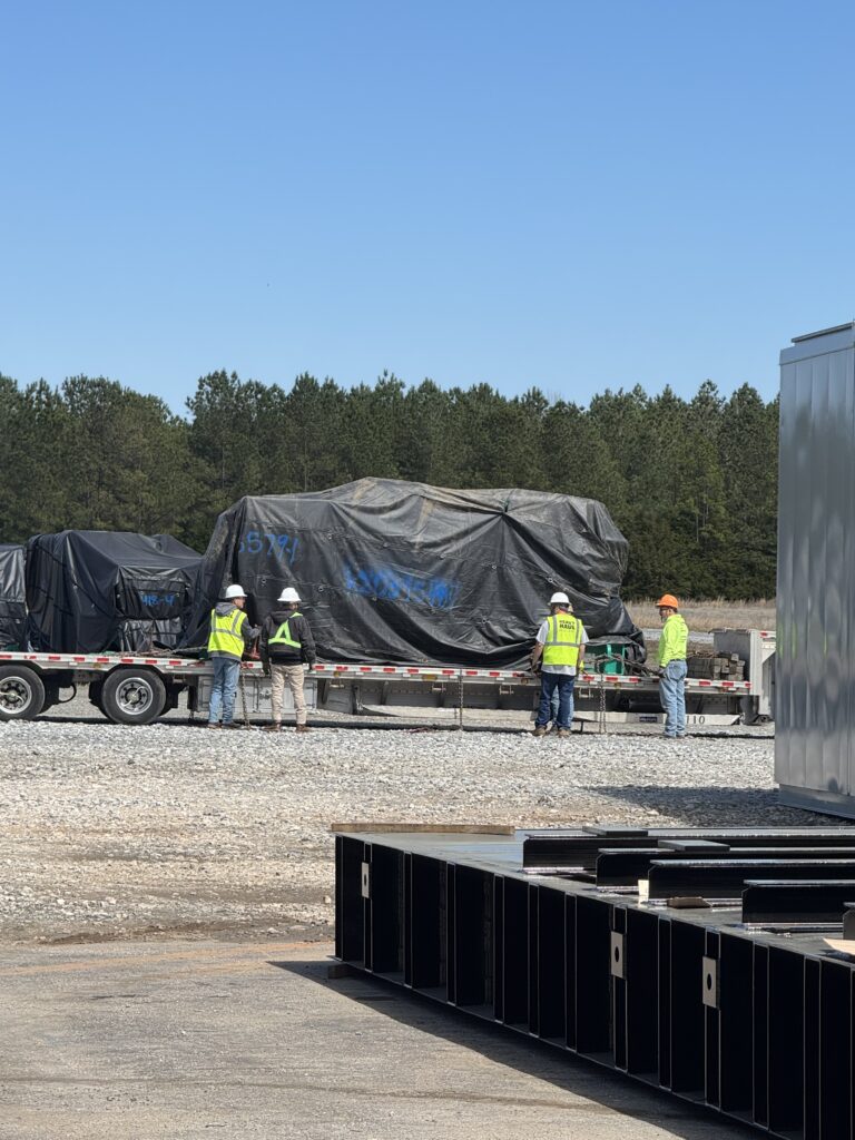 Flatbed trailer parked at yard with oversized cargo covered by black tarp, workers in safety vests nearby, forest backdrop.