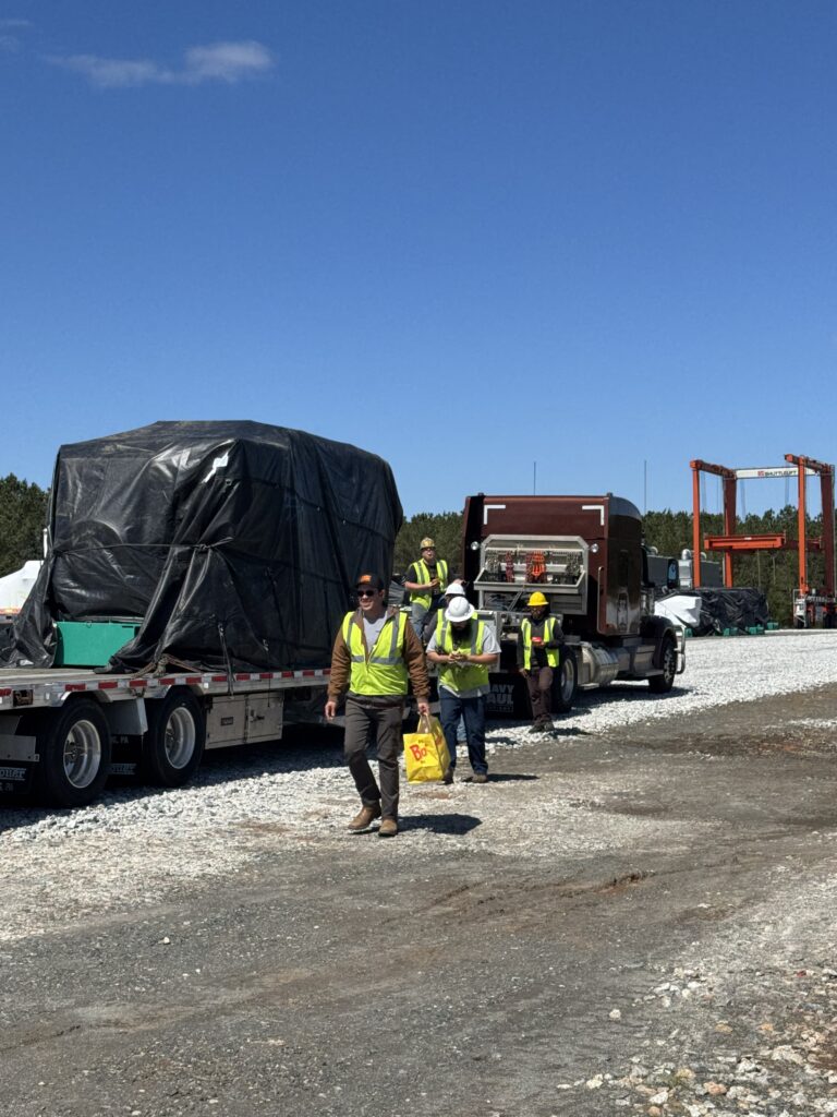Workers in safety vests stand near flatbed trailers and trucks parked at a gravel yard under clear skies.