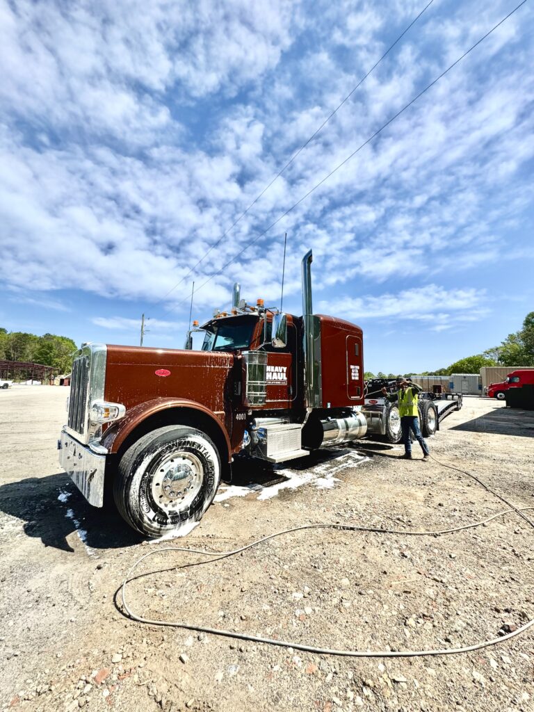 Semi tractor with Heavy Haul Solutions branding parked at a yard; worker in safety vest nearby, with other trucks visible in background.