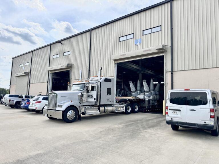 Semi tractor with step deck trailer carrying large industrial equipment parked in front of warehouse facility with other company vehicles nearby