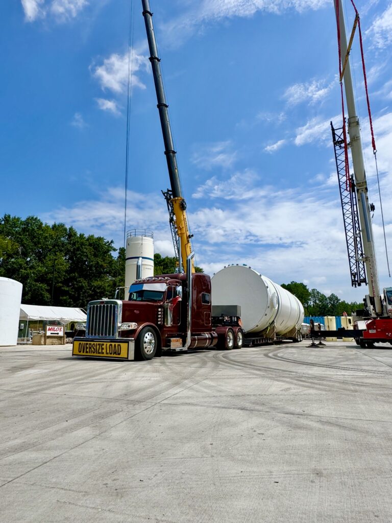 Tanker trailer parked at a yard with two tall cranes and equipment visible in the background under clear sky