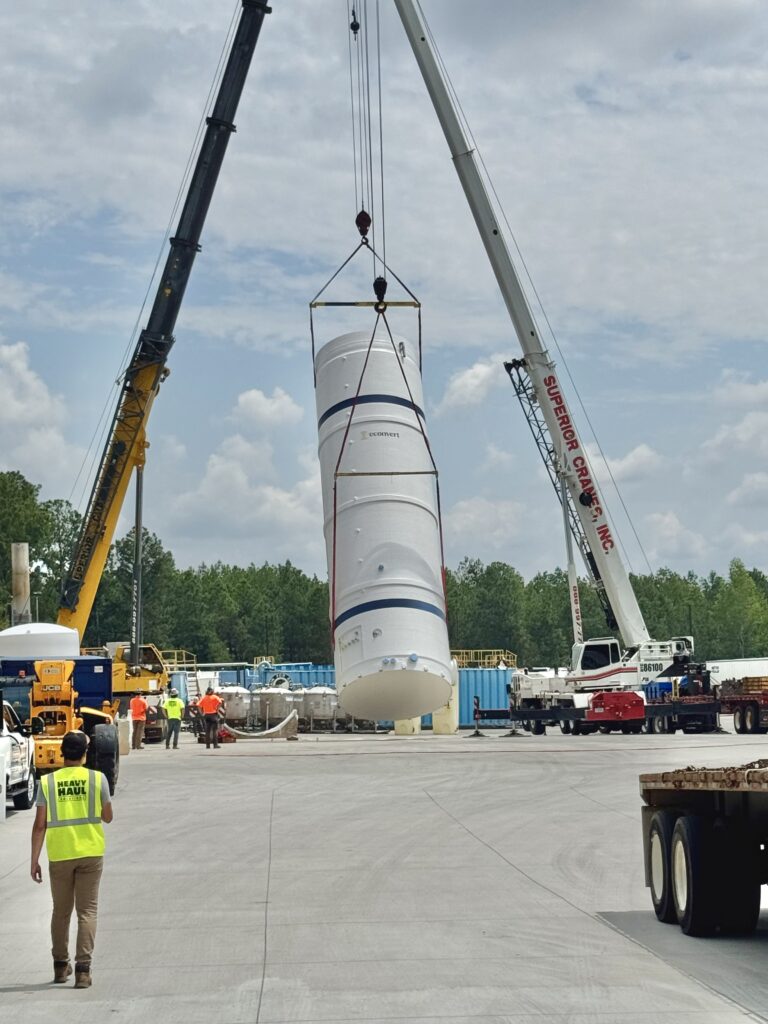 Large cylindrical storage tank suspended by crane rigging between two mobile cranes at an industrial yard with workers in safety gear nearby.