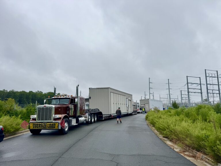 Semi tractor with oversize load sign pulling enclosed trailer down paved road, flagged personnel visible, power transmission structures in background.