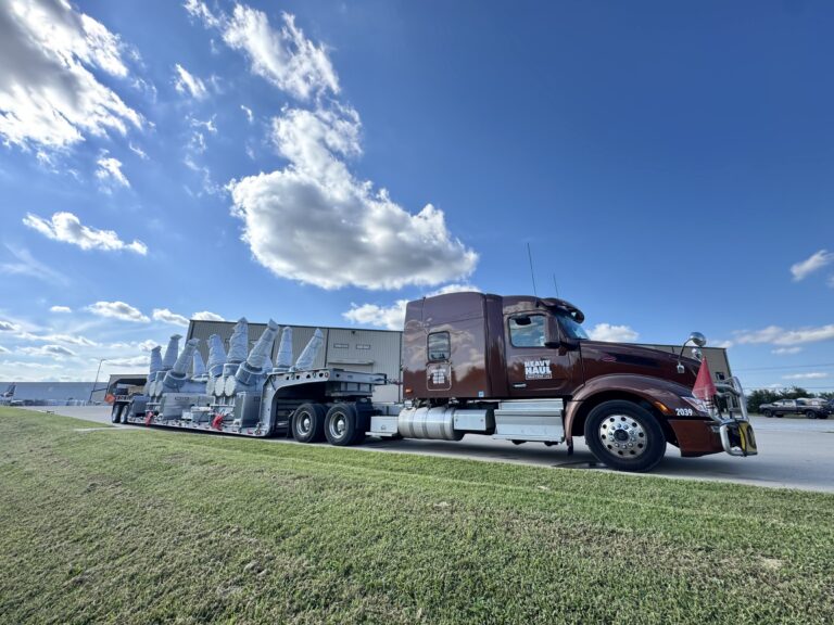 Semi tractor with step deck trailer carrying large industrial modules parked on grass near warehouse buildings under blue sky with clouds.