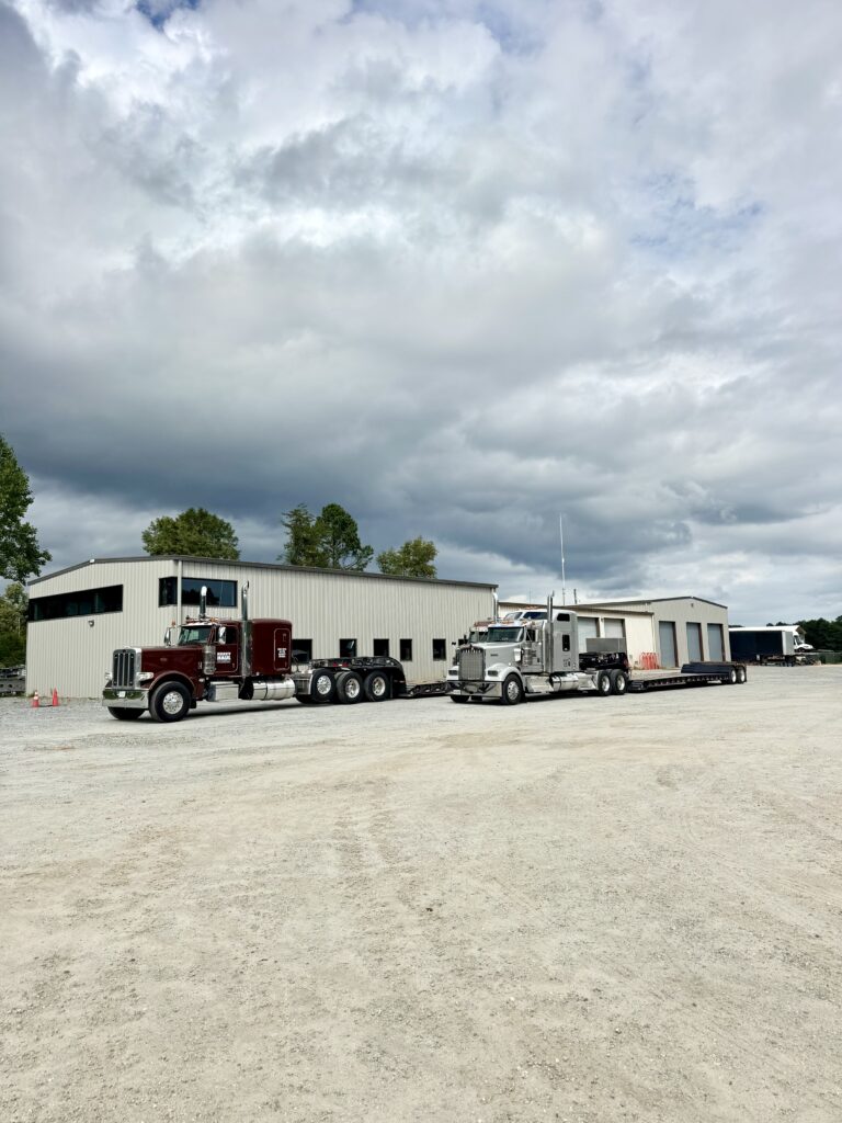 Two semi tractors with enclosed trailers parked at a gravel yard next to industrial buildings under cloudy sky.