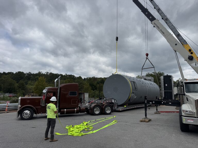 Worker in safety vest guides crane lifting large cylindrical tank onto RGN trailer at industrial yard.