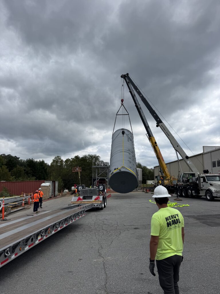 Worker in safety vest watching as crane lifts large cylindrical industrial equipment at job site with overcast sky