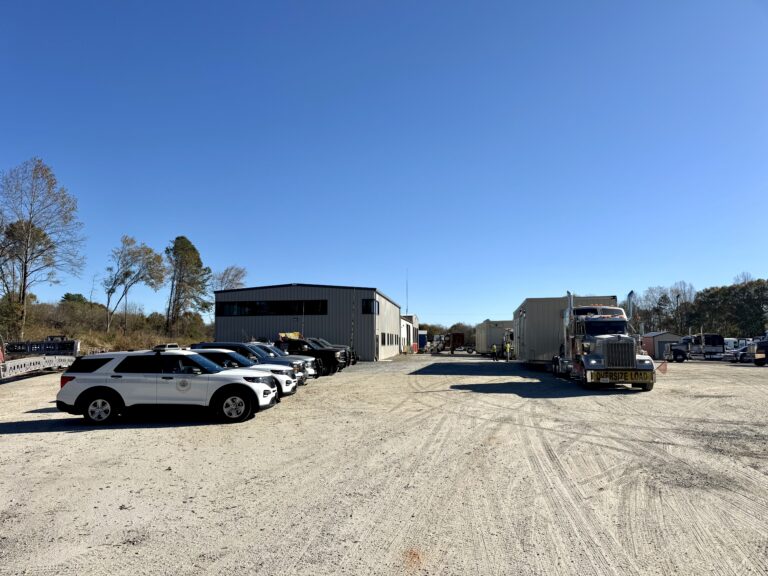 Company yard with parked white pickup trucks and semi tractor with trailer, metal industrial buildings and equipment visible in background under clear sky